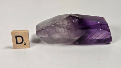 A faceted purple crystal stone, likely amethyst, displayed on a light gray surface next to a small wooden Scrabble tile showing the letter D.