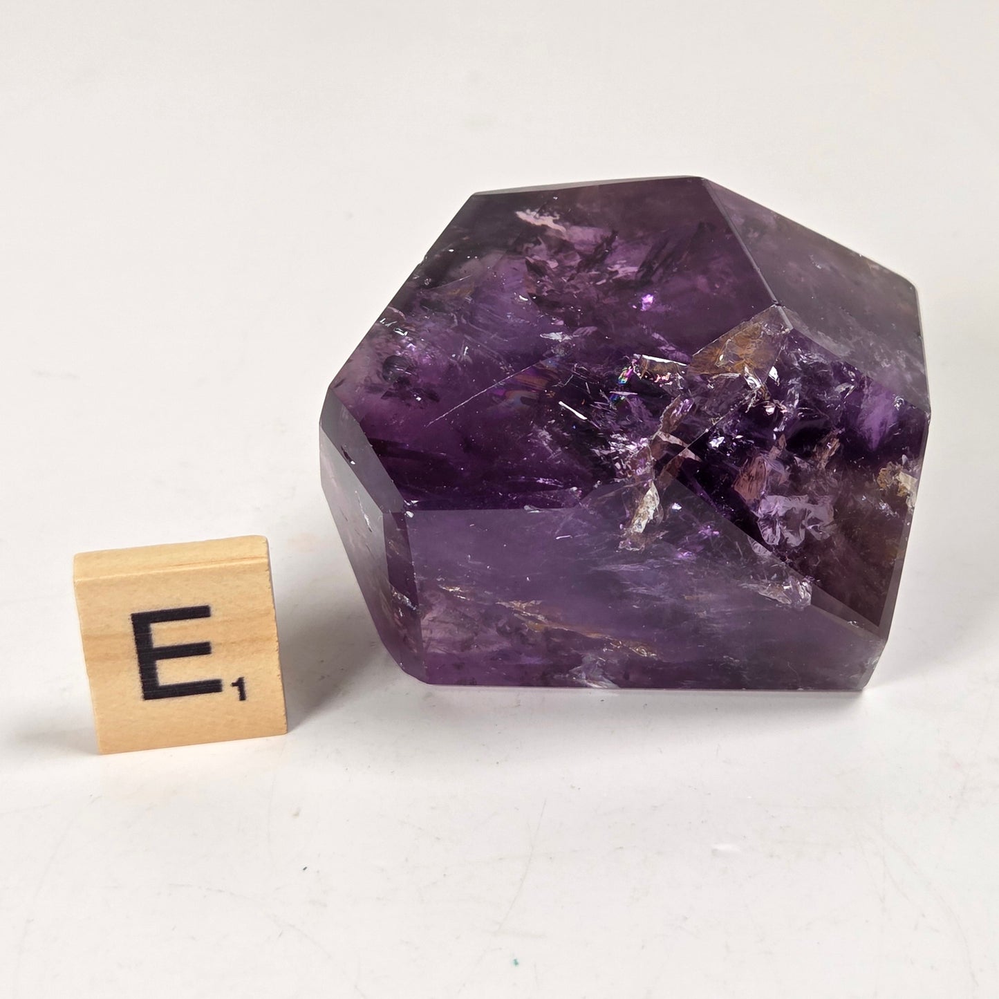 A faceted purple crystal stone, likely amethyst, shown on a light neutral background next to a small wooden Scrabble tile with the letter E.