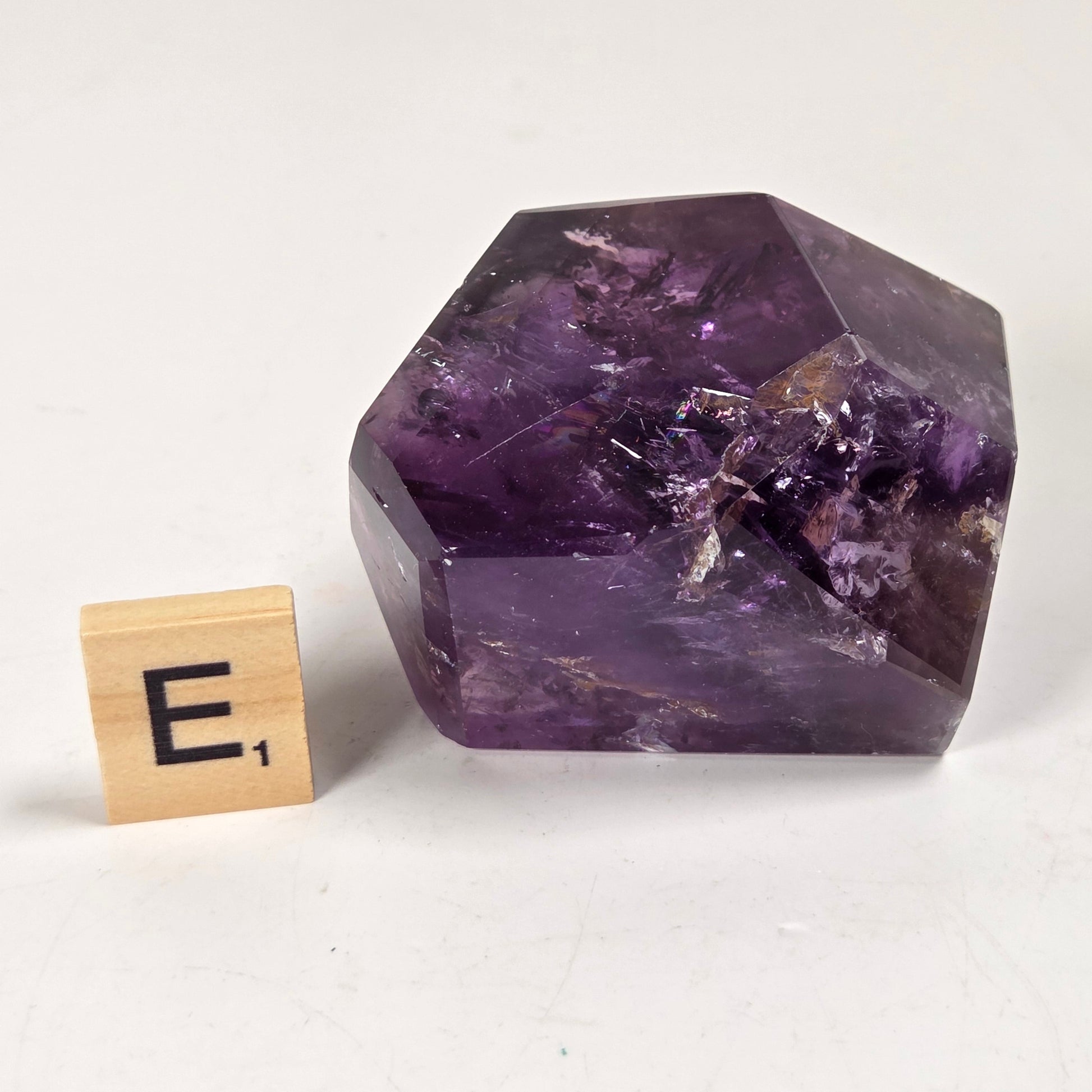 A faceted purple crystal stone, likely amethyst, shown on a light neutral background next to a small wooden Scrabble tile with the letter E.