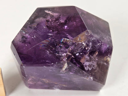 A faceted purple crystal gemstone with natural internal inclusions and a polished, geometric shape, photographed on a plain white background.