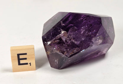 A faceted purple amethyst crystal cluster displayed on a white surface next to a wooden Scrabble tile showing the letter E and its value of 1.
