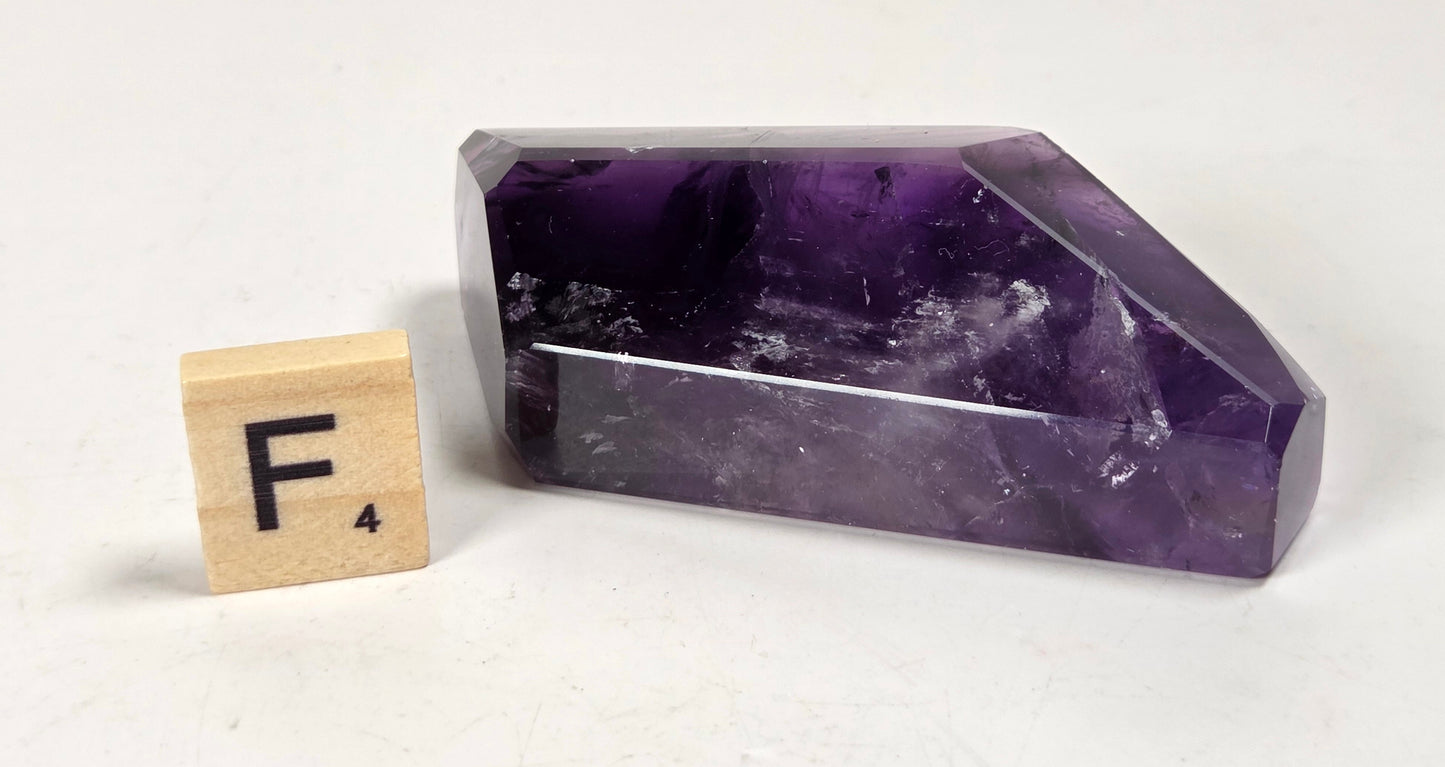 A polished purple amethyst crystal point shown next to a small wooden Scrabble tile with the letter F on a plain off-white background.