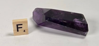 A faceted purple amethyst crystal point displayed on a light gray surface next to a wooden Scrabble tile showing the letter F.