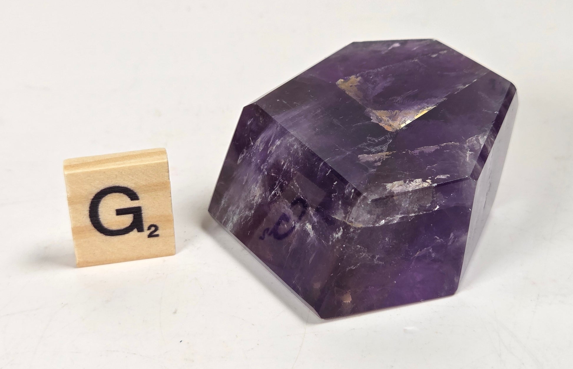 A faceted purple crystal container or box placed on a white surface next to a wooden Scrabble tile showing the letter G.