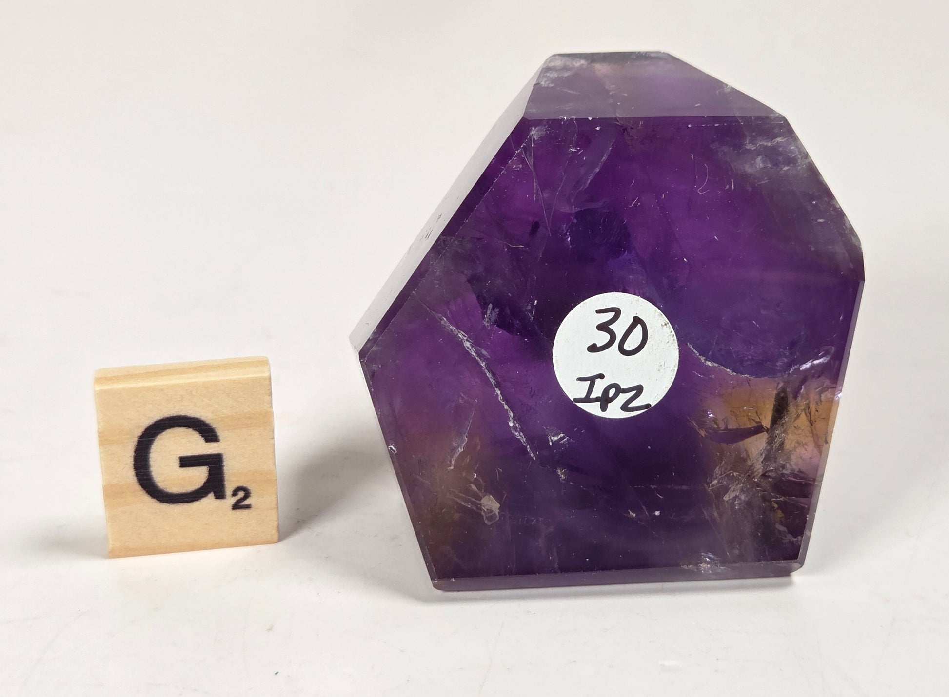 A raw purple amethyst crystal stone with natural inclusions and a white circular sticker reading '30 Joz' next to a small wooden Scrabble tile, set against a plain off-white background.