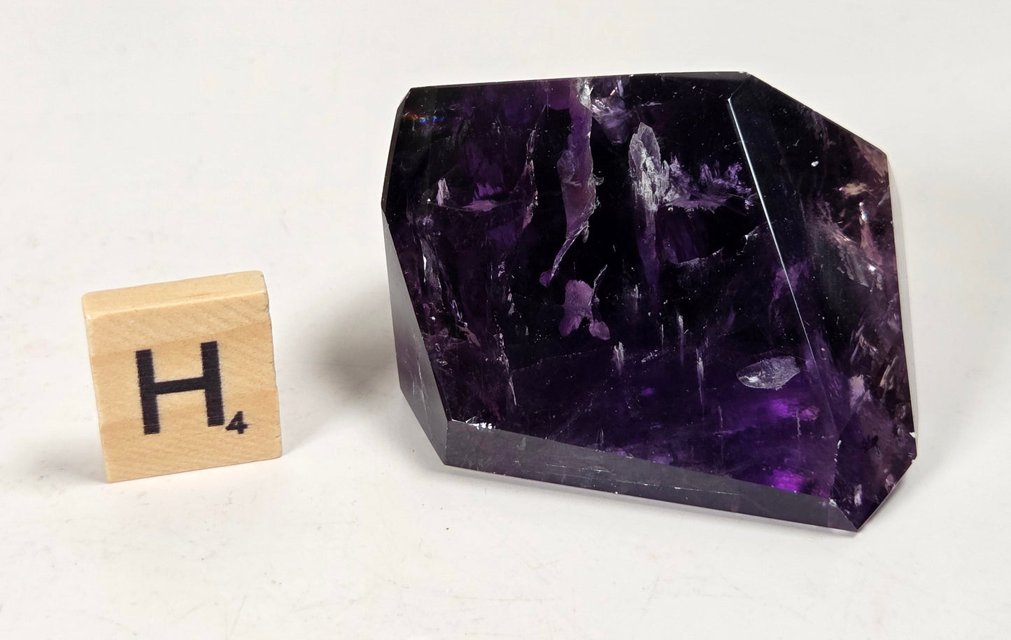 A faceted purple crystal stone with natural inclusions, shown next to a small wooden Scrabble tile displaying the letter H and subscript 4, on a plain off-white background.