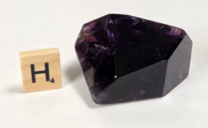 A faceted dark purple crystal stone, likely amethyst, displayed next to a wooden Scrabble tile showing the letter 'H' on a plain white surface.