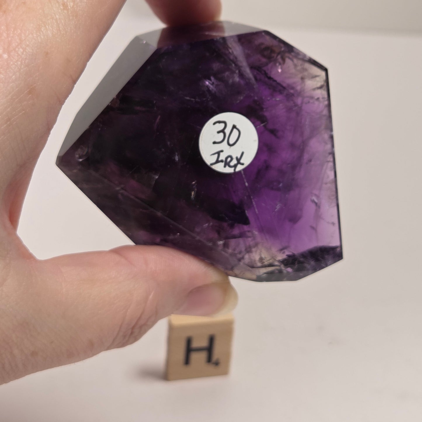 A hand holds a faceted purple amethyst crystal against a plain white background, with a small wooden block displaying the letter 'H' underneath it.