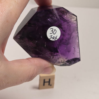 A hand holds a faceted purple amethyst crystal against a plain white background, with a small wooden block displaying the letter 'H' underneath it.