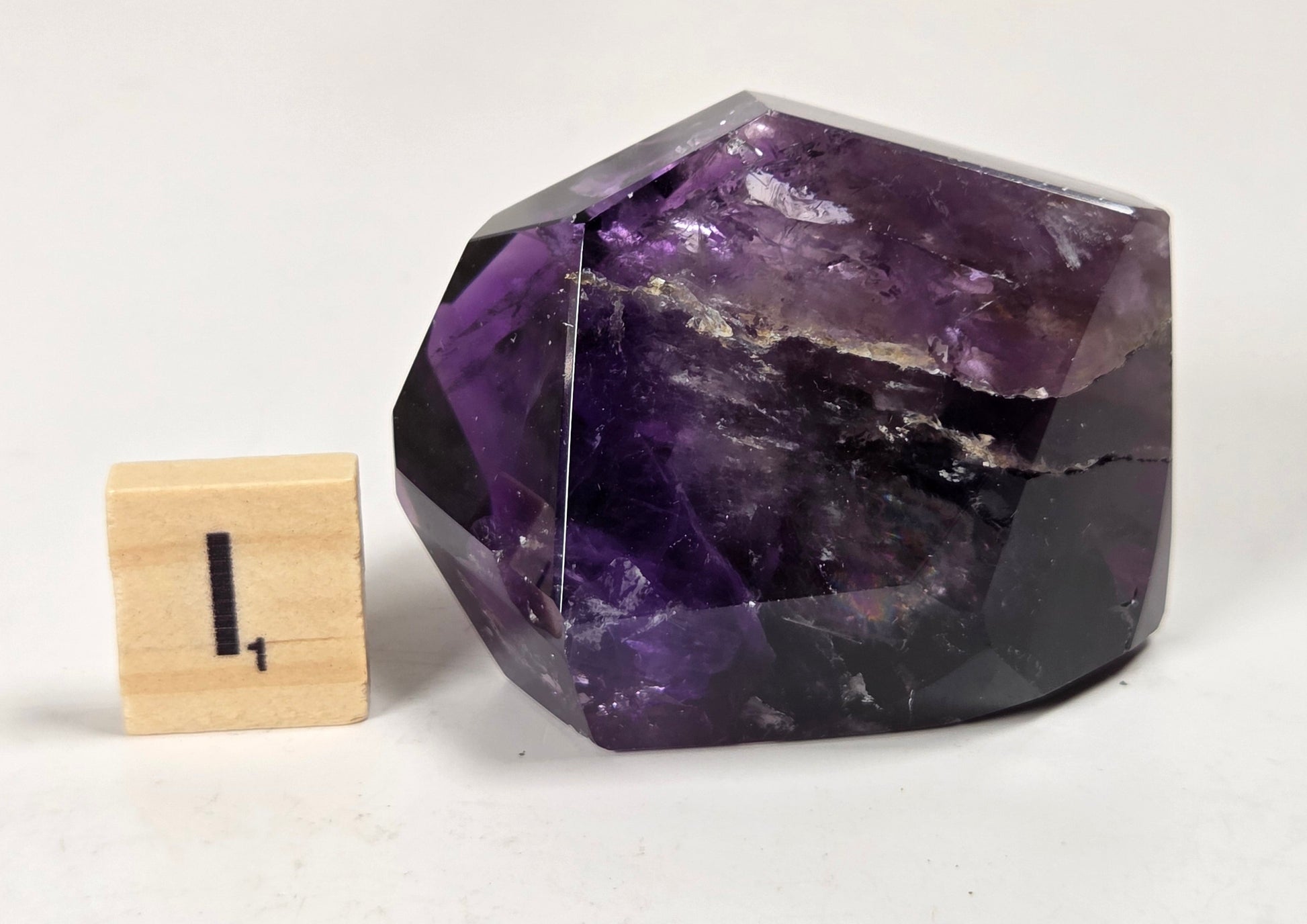 A faceted purple crystal stone, likely amethyst, shown on a white surface next to a small wooden block marked with the number 1 for size reference.