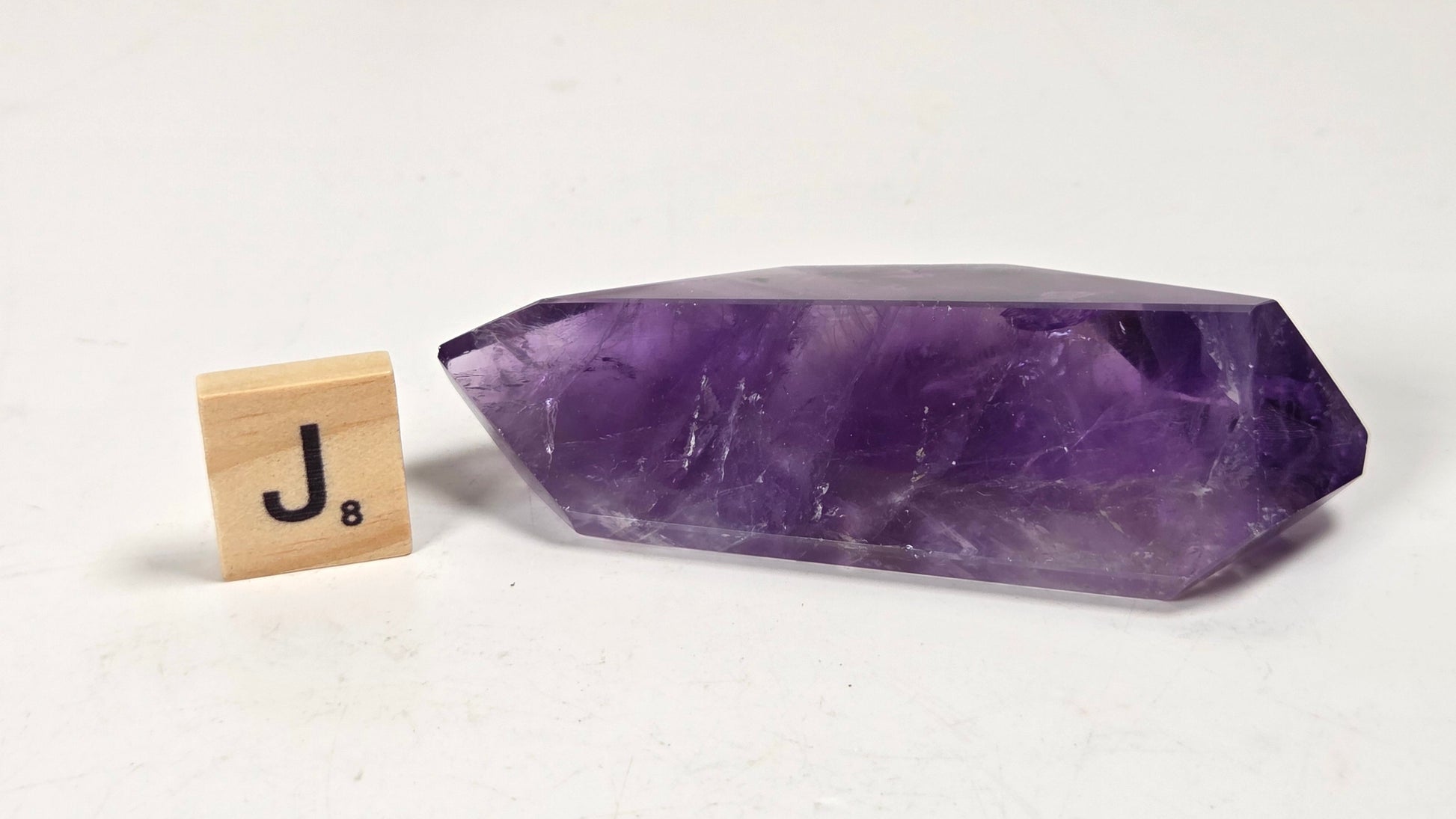 A polished purple crystal point, likely amethyst, displayed on a white surface next to a wooden Scrabble tile showing the letter 'J'.