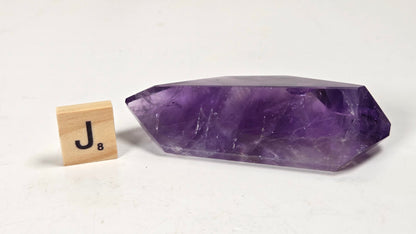 A polished purple crystal point, likely amethyst, displayed on a white surface next to a wooden Scrabble tile showing the letter 'J'.