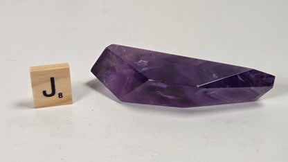 A faceted purple crystal mineral specimen displayed on a light gray surface next to a small wooden Scrabble tile showing the letter J.