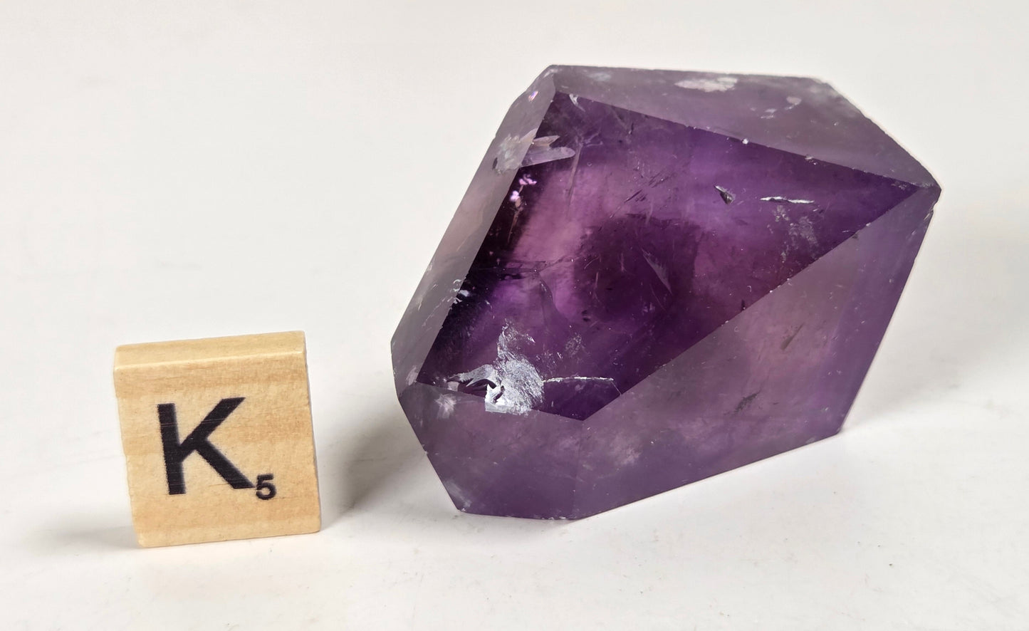 A faceted purple amethyst crystal stone displayed on a white surface next to a wooden Scrabble tile showing the letter K and the score 5.
