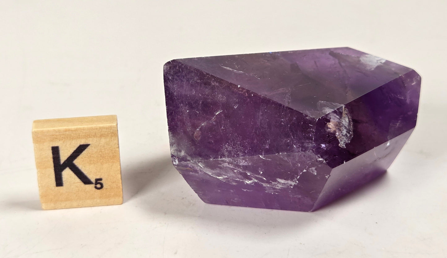 A faceted purple amethyst crystal stone shown next to a wooden Scrabble tile spelling 'K' on a plain off-white background.