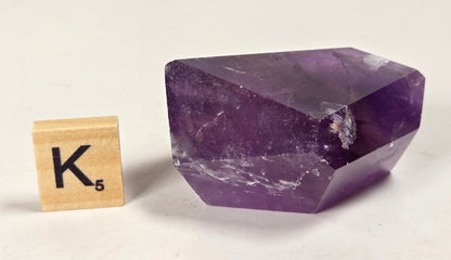 A faceted purple amethyst crystal stone shown next to a wooden Scrabble tile spelling 'K' on a plain off-white background.