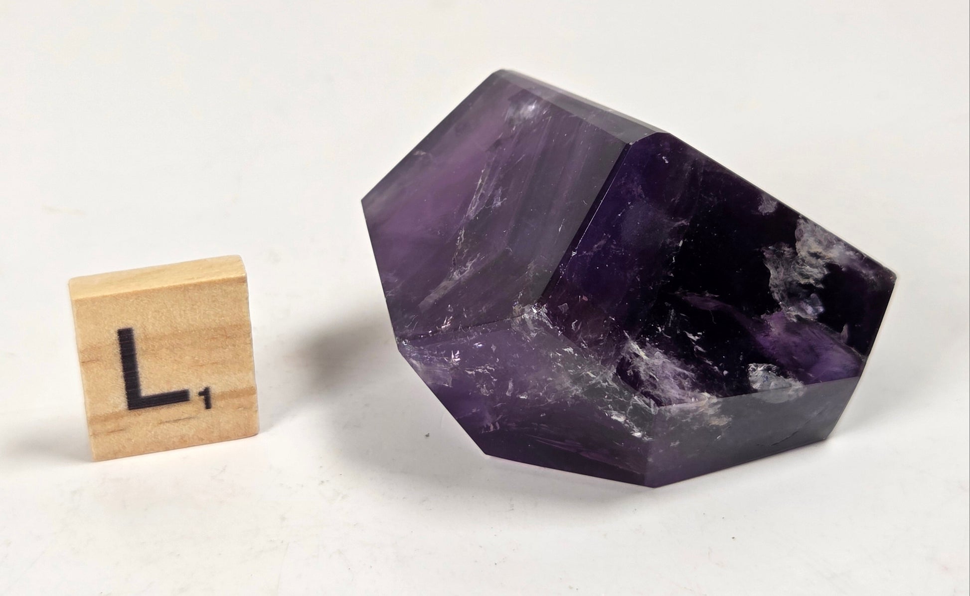 A polished purple amethyst crystal stone displayed next to a small wooden letter tile with the letter 'L' on a plain white background.