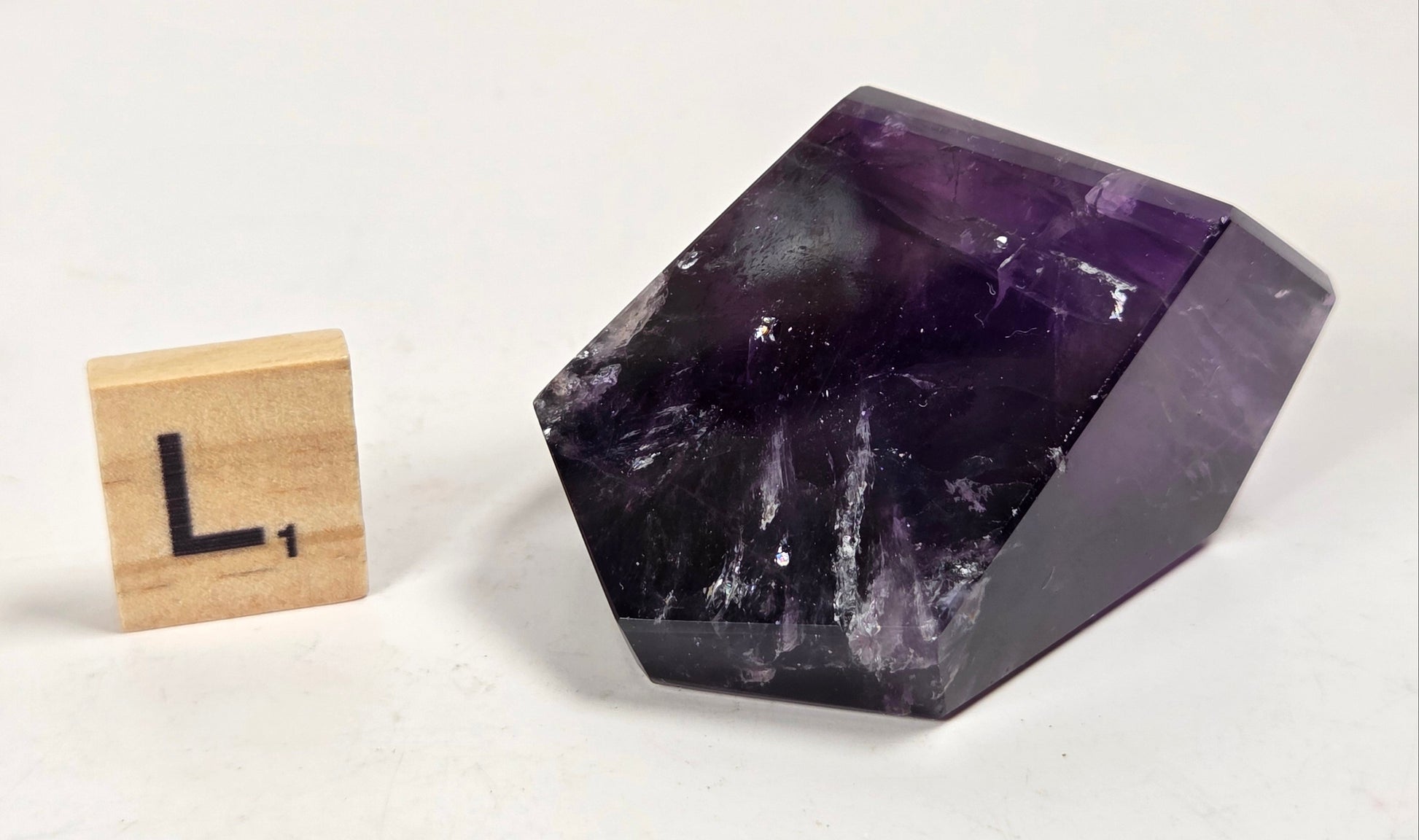 A polished purple amethyst crystal stone with natural white and dark veining, shown next to a wooden Scrabble tile with the letter L on a plain off-white background.