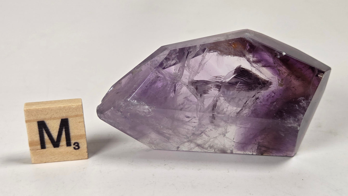 A raw amethyst crystal point with purple and lavender tones, shown next to a wooden Scrabble tile spelling 'M3' on a light gray background.