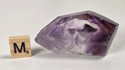 A raw amethyst crystal point with purple and lavender tones, shown next to a wooden Scrabble tile spelling 'M3' on a light gray background.