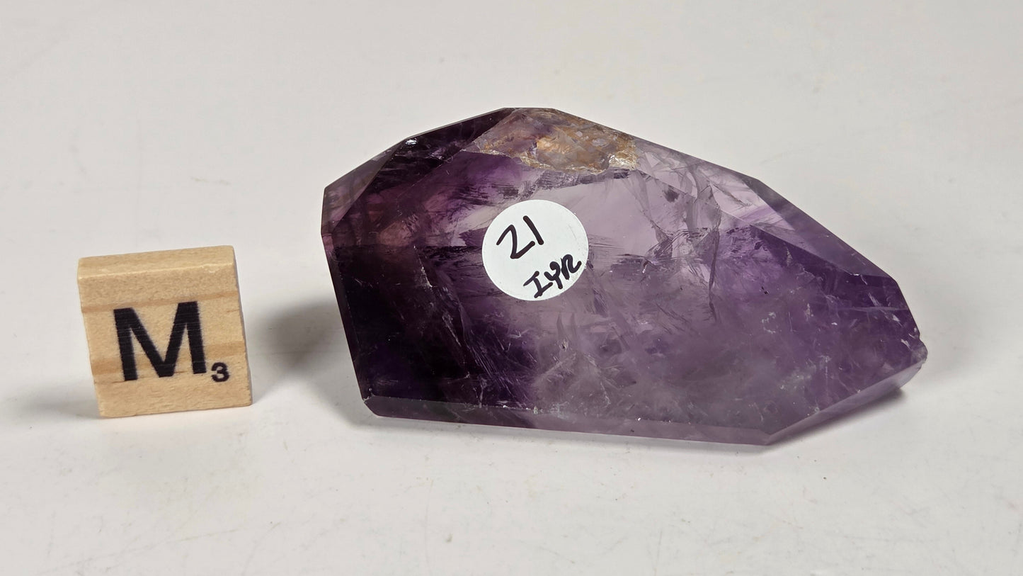 A raw purple amethyst crystal point with a white circular sticker reading '21 JSPC' next to a wooden Scrabble tile showing 'M3', photographed on a light gray surface.