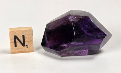 A faceted purple crystal gemstone displayed next to a wooden Scrabble tile showing the letter 'N', set against a plain off-white background.