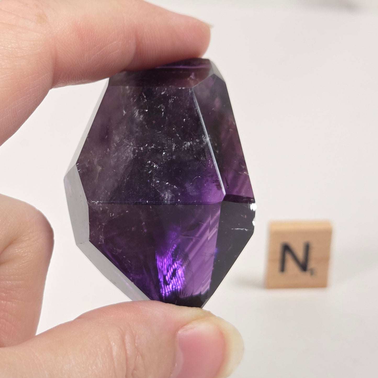 A hand holding a faceted purple crystal gemstone, with a small Scrabble tile in the background on a white surface.