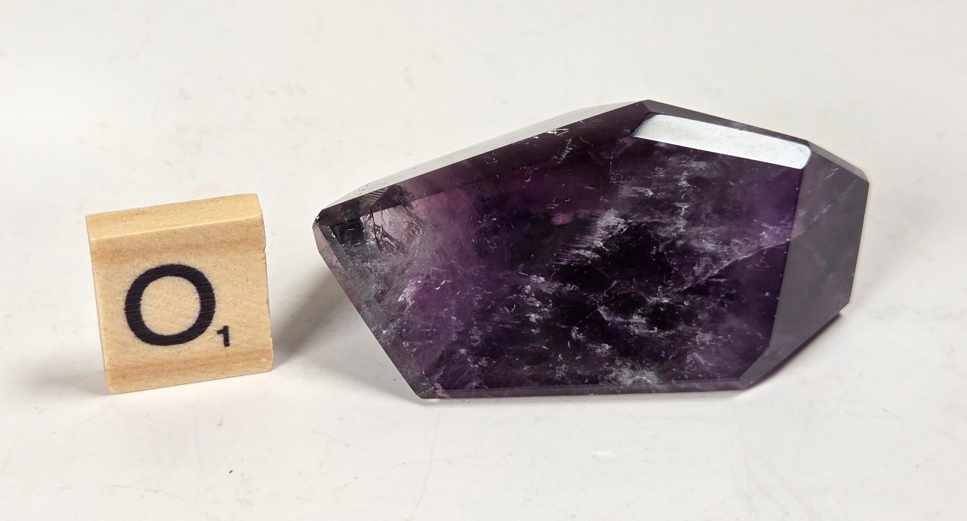 A faceted purple amethyst crystal stone displayed next to a wooden Scrabble tile showing the letter 'O' on a light off-white background.
