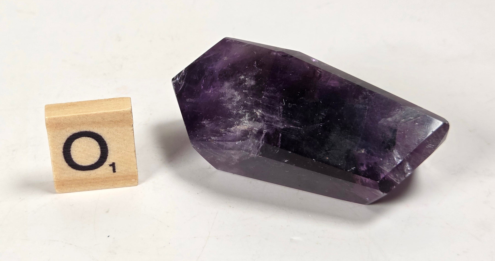 A polished purple amethyst crystal point shown next to a small wooden Scrabble tile on a plain off-white background.