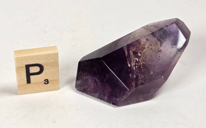A faceted purple amethyst crystal stone displayed next to a wooden Scrabble tile showing the letter P and value 3, set against a plain off-white background.