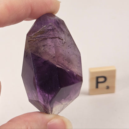 A hand holds a faceted purple amethyst crystal point against a plain white background, with a small wooden Scrabble tile in the background.