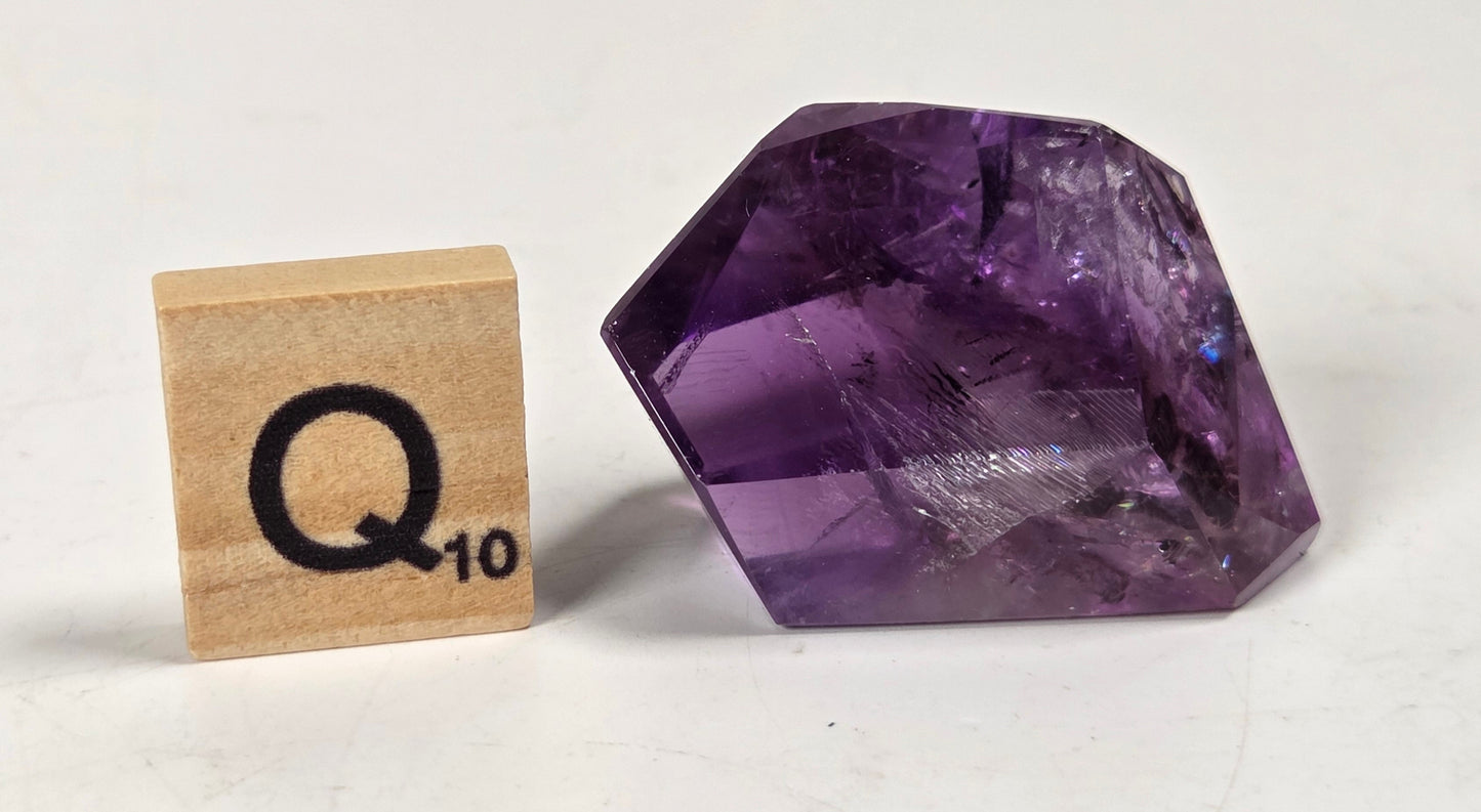 A faceted purple crystal gemstone shown next to a wooden Scrabble tile marked 'Q 10' on a plain off-white surface.