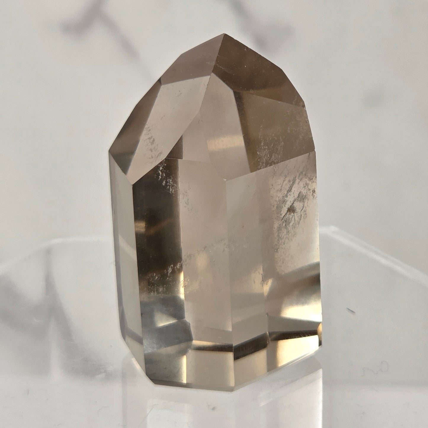 A faceted smoky quartz crystal point displayed on a clear acrylic stand against a light marble-patterned background.