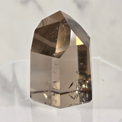 A natural clear quartz crystal point with internal inclusions, displayed on a clear acrylic stand against a light gray marble background.