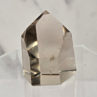 A natural clear quartz crystal point with internal inclusions, displayed on a clear acrylic stand against a light gray marble background.