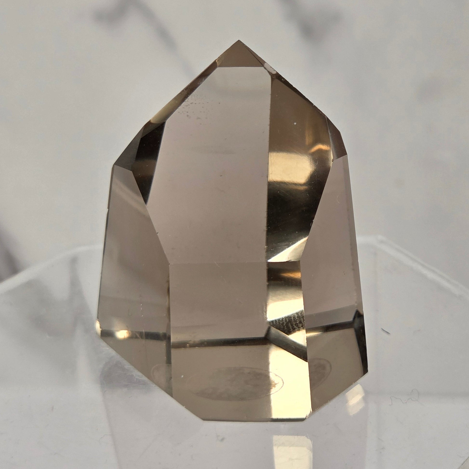 A faceted smoky quartz crystal display piece resting on a clear acrylic stand against a light gray marble-patterned background.