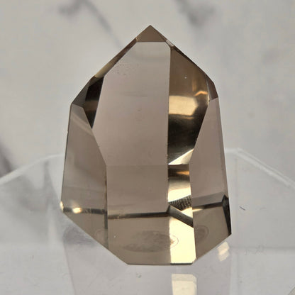 A faceted smoky quartz crystal display piece resting on a clear acrylic stand against a light gray marble-patterned background.