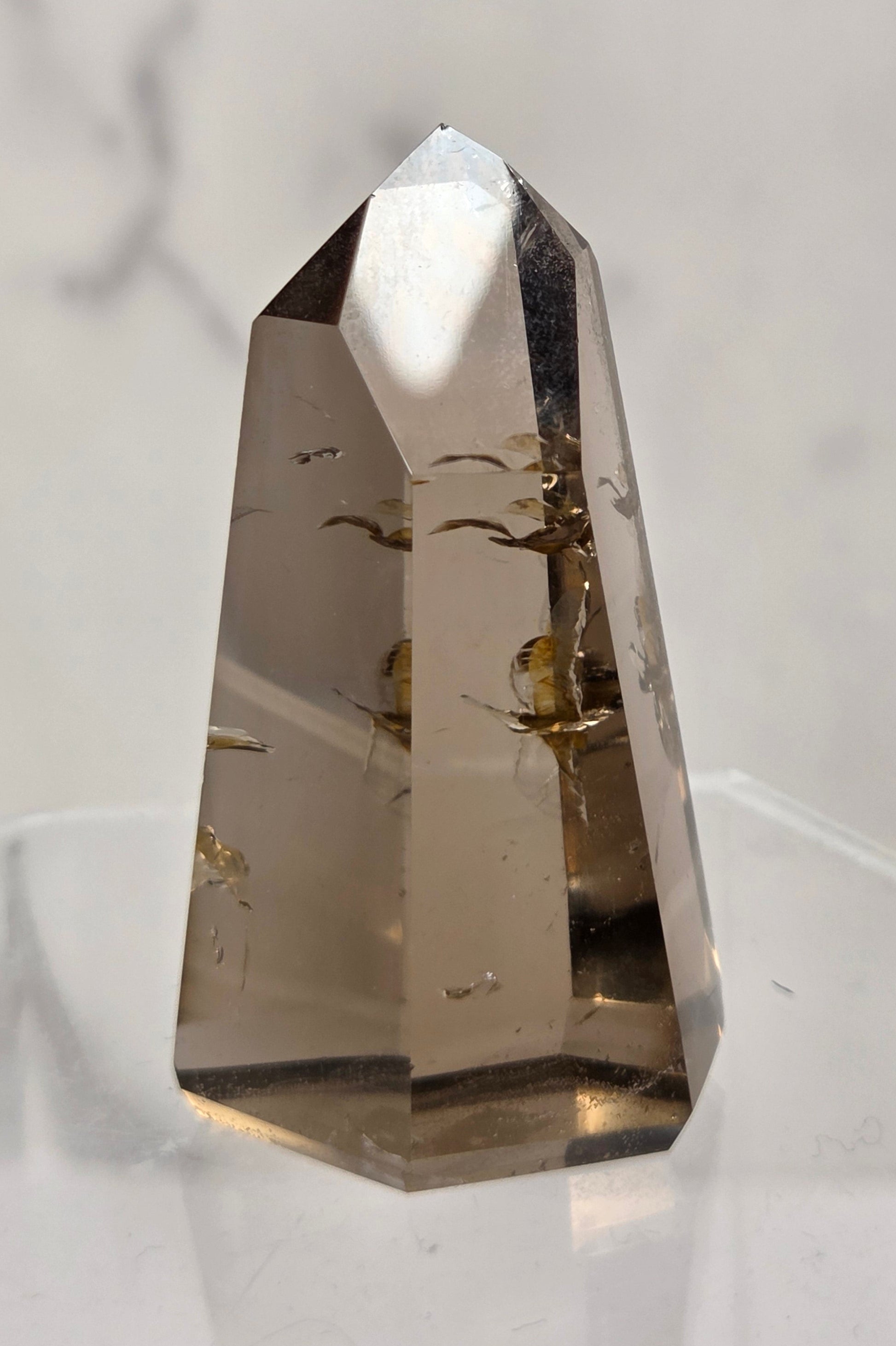 A natural smoky quartz crystal point with inclusions, displayed on a clear acrylic stand against a light marble background.