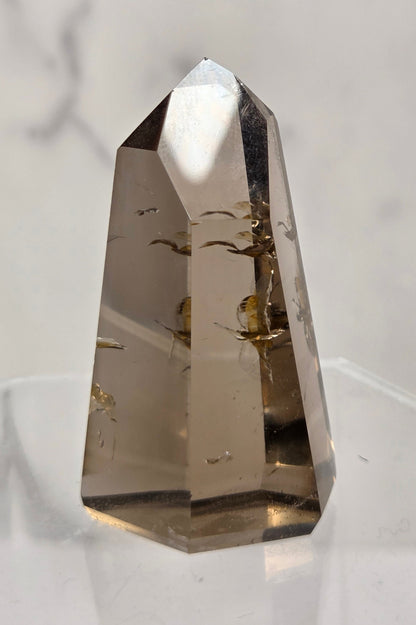 A natural smoky quartz crystal point with inclusions, displayed on a clear acrylic stand against a light marble background.