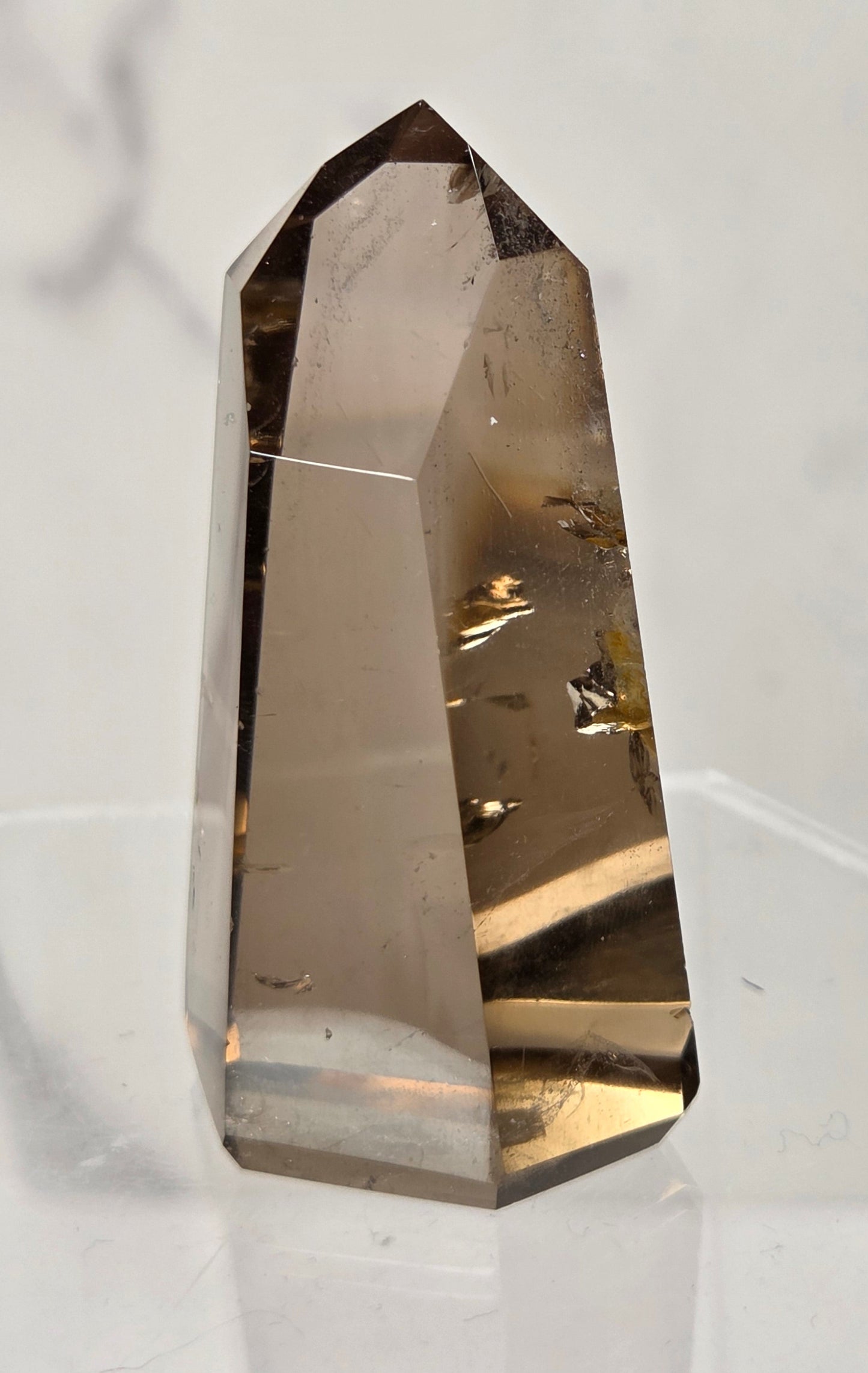 A faceted smoky quartz crystal point with natural inclusions, displayed on a clear acrylic stand against a light gray marble-patterned background.