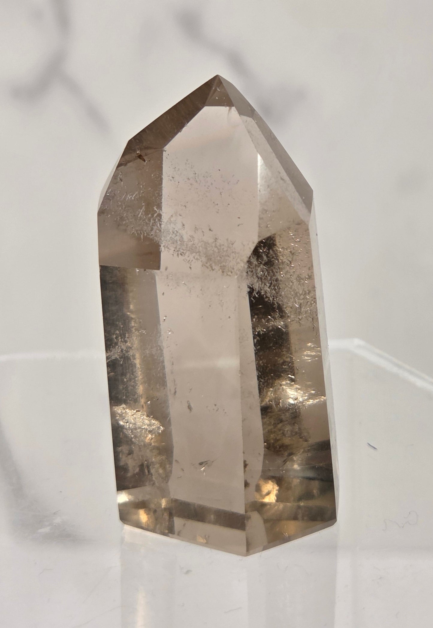 A natural smoky quartz crystal point with internal inclusions, displayed on a clear acrylic stand against a light gray marble-patterned background.