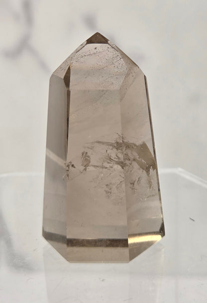 A natural smoky quartz crystal point with internal inclusions, displayed on a clear acrylic stand against a light marble background.