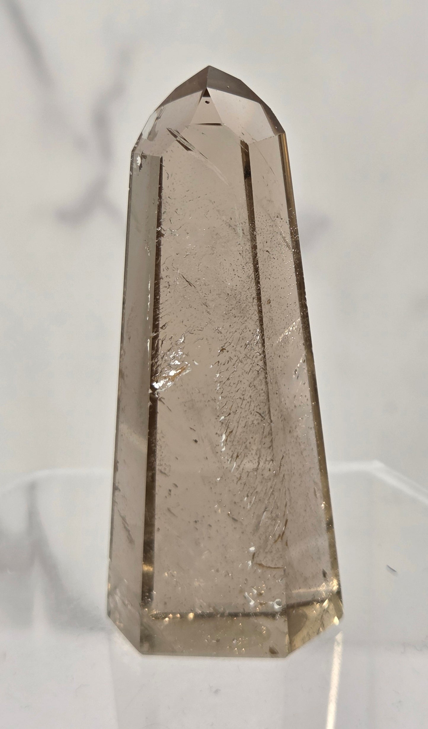A faceted smoky quartz crystal tower on a clear acrylic display stand, set against a light gray marble-patterned background.