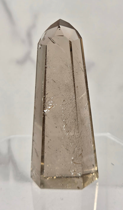 A faceted smoky quartz crystal tower on a clear acrylic display stand, set against a light gray marble-patterned background.