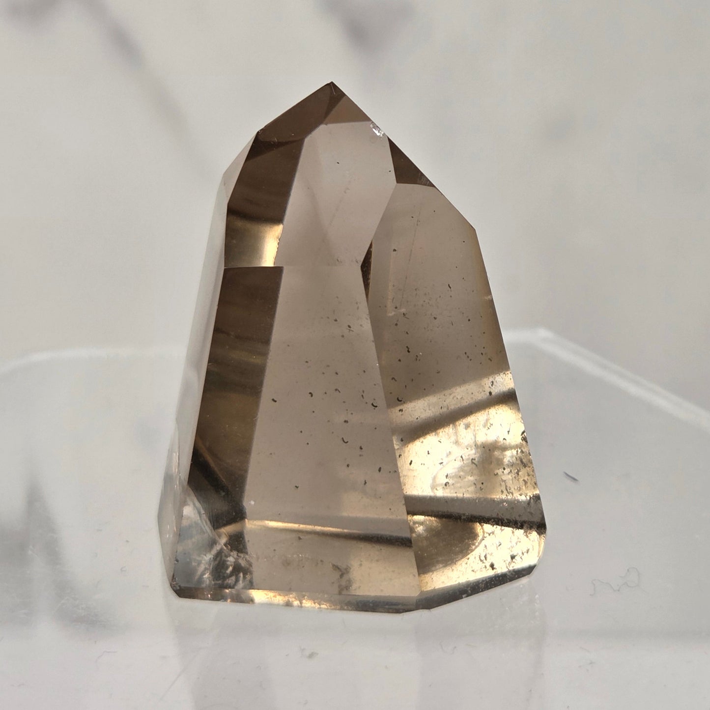 A natural smoky quartz crystal point displayed on a clear acrylic stand against a light gray marble-patterned background.