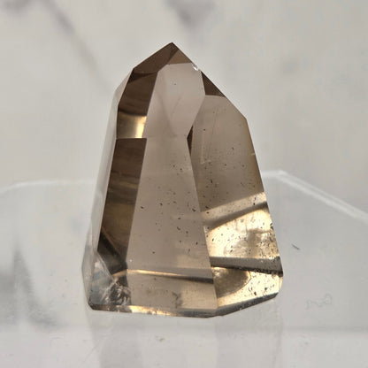 A natural smoky quartz crystal point displayed on a clear acrylic stand against a light gray marble-patterned background.