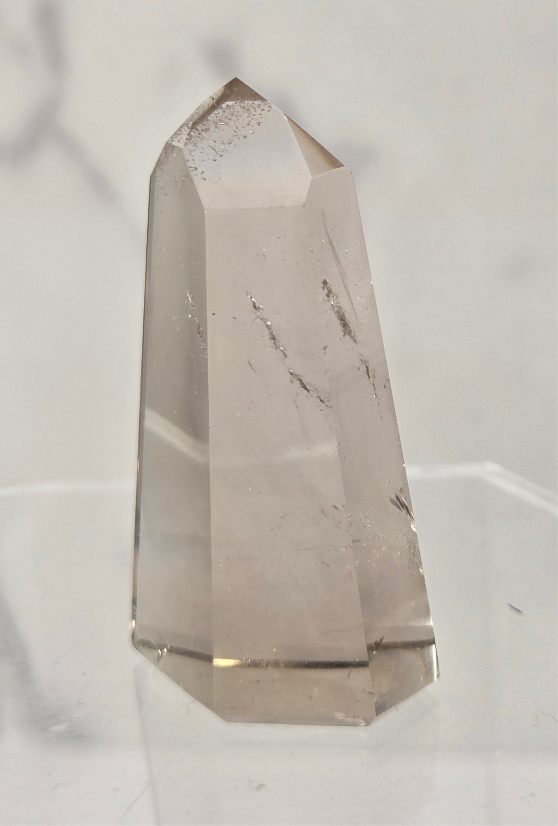 A natural smoky quartz crystal tower with internal inclusions, displayed on a clear acrylic stand against a light gray marble-patterned background.