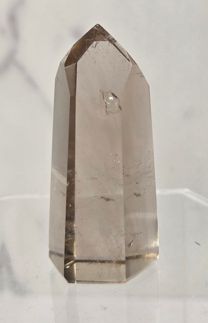 A natural smoky quartz crystal point tower displayed on a clear acrylic stand against a light gray marble-patterned background.