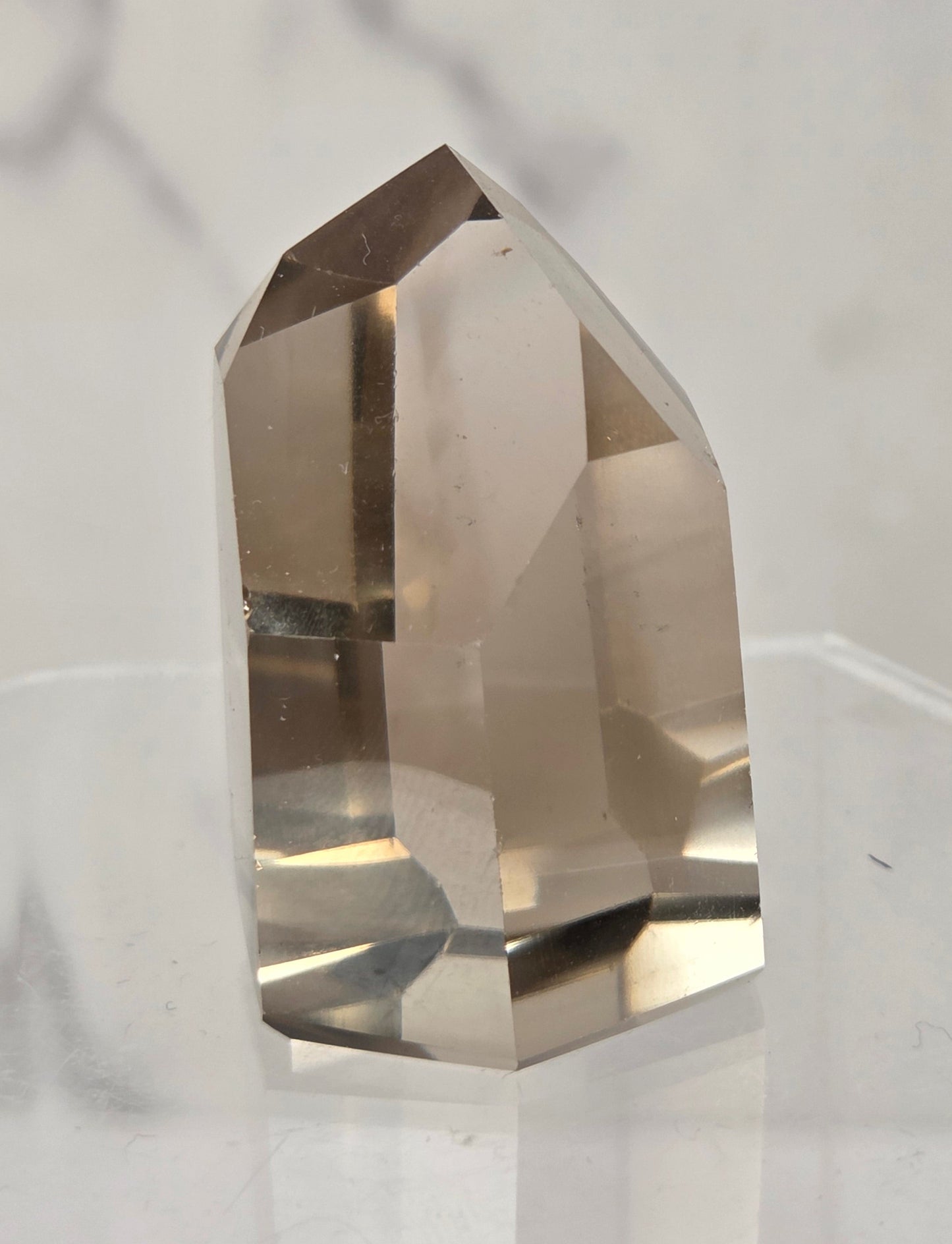 A natural smoky quartz crystal point with a hexagonal shape, standing upright on a clear acrylic display block against a light gray marble background.