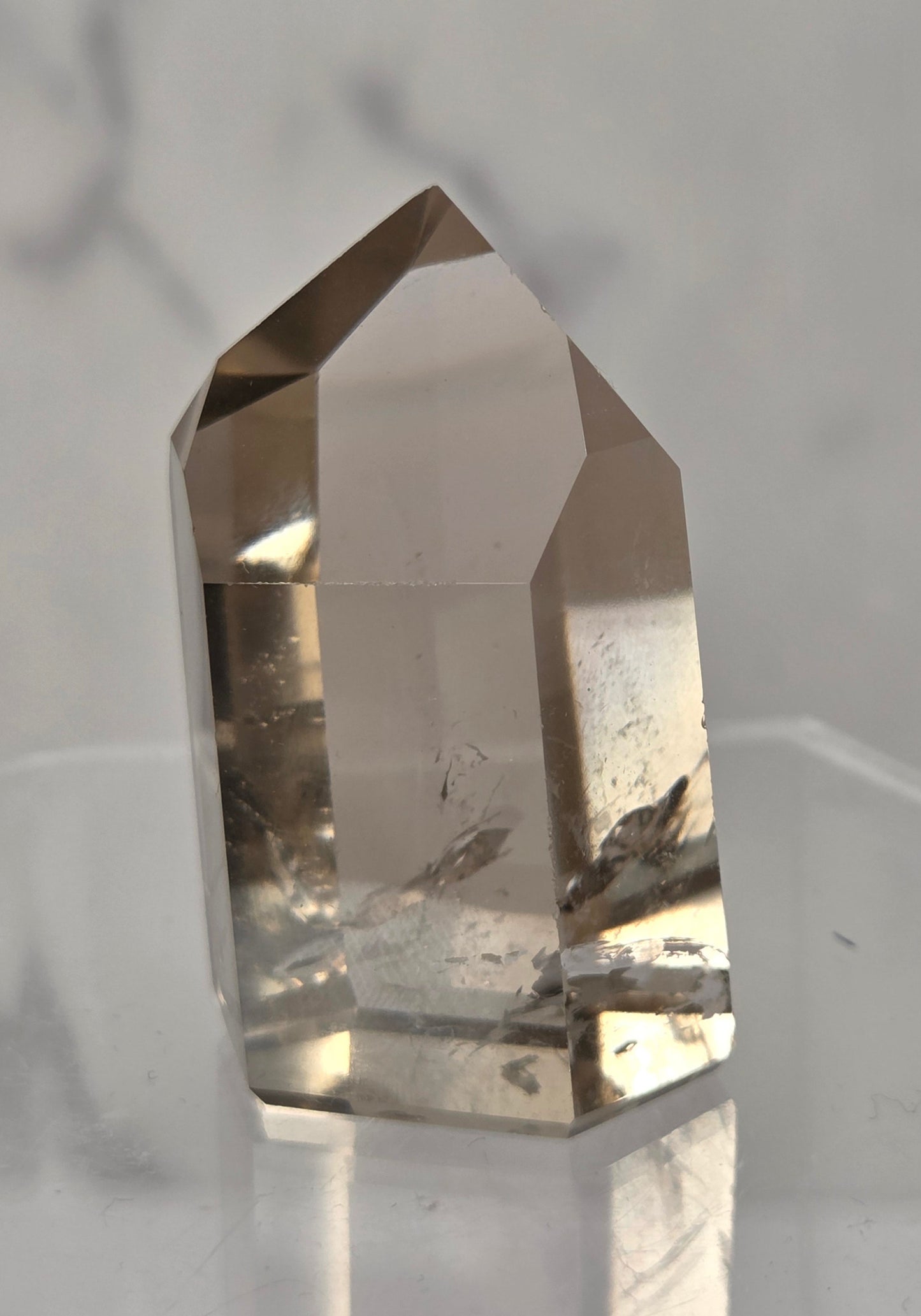 A natural smoky quartz crystal point with internal inclusions, displayed on a clear acrylic stand against a light gray marble background.